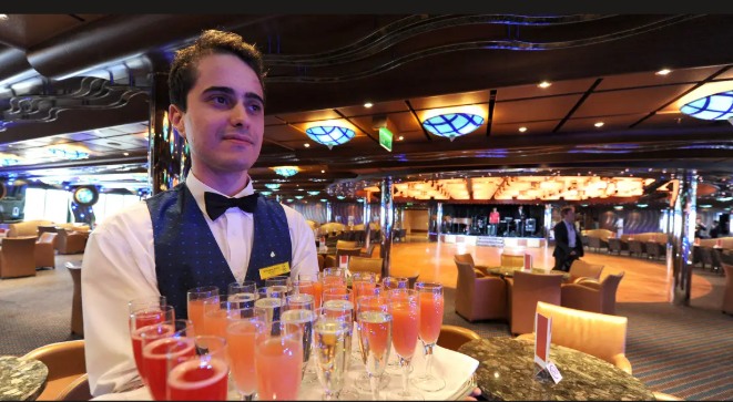 A beverage server carrying a tray of drinks through the ship's lounge, tables and seating visible across the room